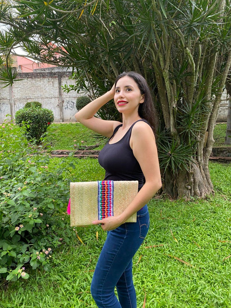 Handmade palm clutch featuring a rectangular woven palm leaf body, decorated with vibrant multicolor tassels and hand-painted pine cone flowers in pink, yellow, turquoise, purple, orange, and red. The center strip has a colorful embroidered textile accent, creating a unique artisan boho design. Perfect as a statement accessory for festivals, summer parties, vacations, or as a one-of-a-kind handmade gift for women.
