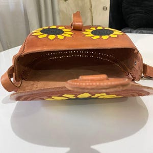 Handcrafted Mexican leather crossbody bag modeled by a woman in a rust sleeveless top and denim jeans. The bag is tan brown with hand-tooled sunflower details in bright yellow and black, featuring a curved flap, rustic stitching, and an adjustable leather strap. A unique bohemian accessory perfect for adding handmade floral charm and artisan craftsmanship to everyday outfits or travel looks.