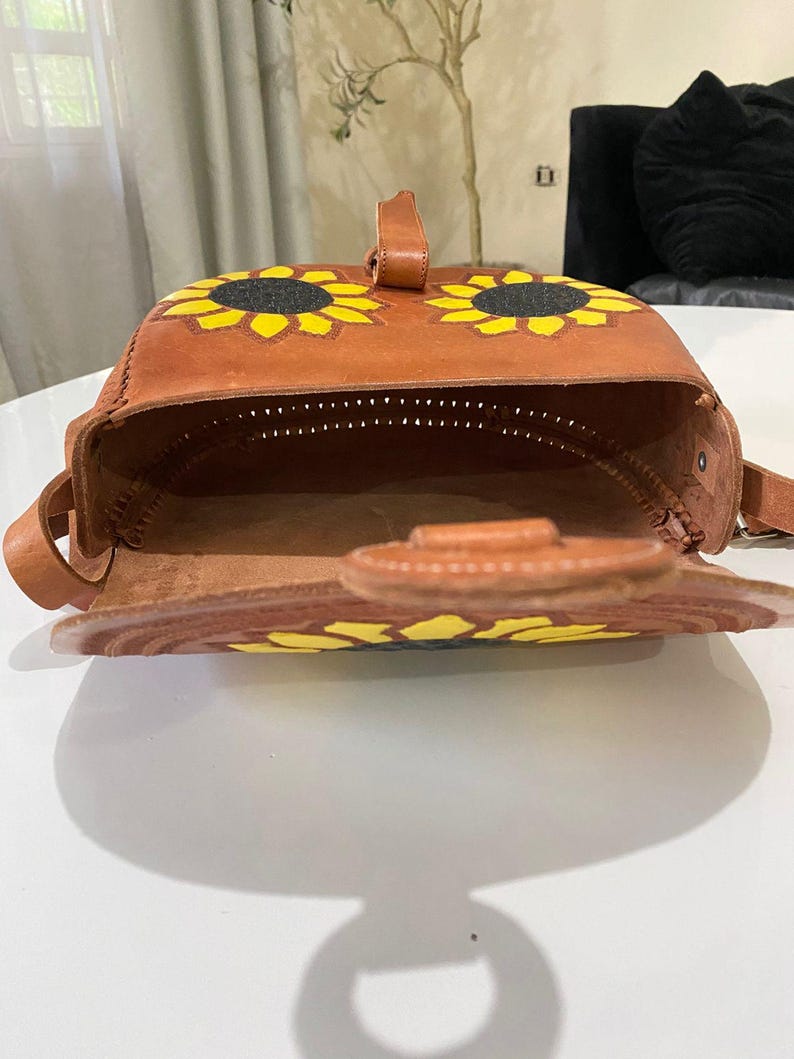 Handcrafted Mexican leather crossbody bag modeled by a woman in a rust sleeveless top and denim jeans. The bag is tan brown with hand-tooled sunflower details in bright yellow and black, featuring a curved flap, rustic stitching, and an adjustable leather strap. A unique bohemian accessory perfect for adding handmade floral charm and artisan craftsmanship to everyday outfits or travel looks.
