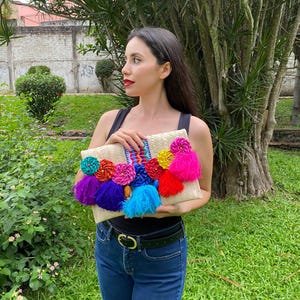 Handmade palm clutch featuring a rectangular woven palm leaf body, decorated with vibrant multicolor tassels and hand-painted pine cone flowers in pink, yellow, turquoise, purple, orange, and red. The center strip has a colorful embroidered textile accent, creating a unique artisan boho design. Perfect as a statement accessory for festivals, summer parties, vacations, or as a one-of-a-kind handmade gift for women.