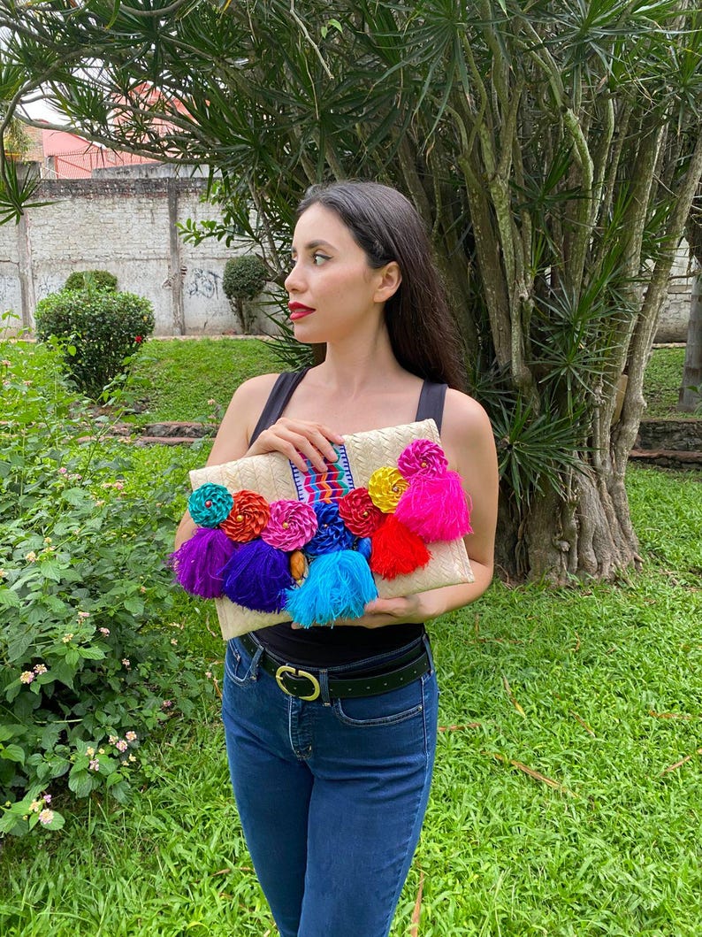 Handmade palm clutch featuring a rectangular woven palm leaf body, decorated with vibrant multicolor tassels and hand-painted pine cone flowers in pink, yellow, turquoise, purple, orange, and red. The center strip has a colorful embroidered textile accent, creating a unique artisan boho design. Perfect as a statement accessory for festivals, summer parties, vacations, or as a one-of-a-kind handmade gift for women.