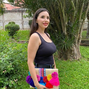 Handmade palm clutch featuring a rectangular woven palm leaf body, decorated with vibrant multicolor tassels and hand-painted pine cone flowers in pink, yellow, turquoise, purple, orange, and red. The center strip has a colorful embroidered textile accent, creating a unique artisan boho design. Perfect as a statement accessory for festivals, summer parties, vacations, or as a one-of-a-kind handmade gift for women.