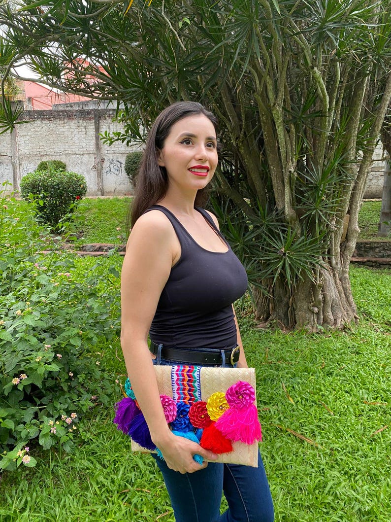Handmade palm clutch featuring a rectangular woven palm leaf body, decorated with vibrant multicolor tassels and hand-painted pine cone flowers in pink, yellow, turquoise, purple, orange, and red. The center strip has a colorful embroidered textile accent, creating a unique artisan boho design. Perfect as a statement accessory for festivals, summer parties, vacations, or as a one-of-a-kind handmade gift for women.