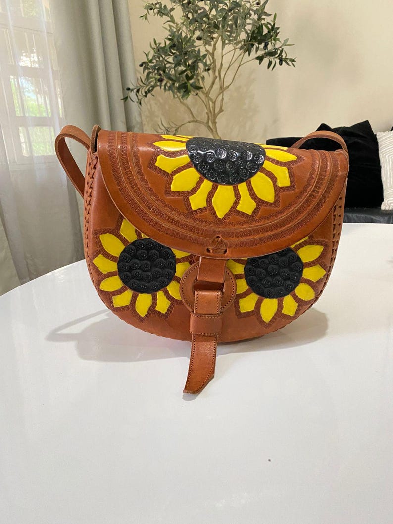 Handcrafted Mexican leather crossbody bag modeled by a woman in a rust sleeveless top and denim jeans. The bag is tan brown with hand-tooled sunflower details in bright yellow and black, featuring a curved flap, rustic stitching, and an adjustable leather strap. A unique bohemian accessory perfect for adding handmade floral charm and artisan craftsmanship to everyday outfits or travel looks.