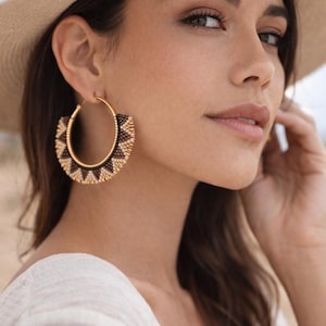 May include: Gold hoop earrings with a geometric pattern in black and tan beads. The earrings are large and eye-catching, with a circular design. The model is wearing a cream-colored top and a beige hat.