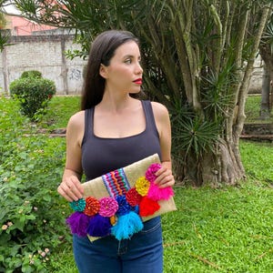 Handmade palm clutch featuring a rectangular woven palm leaf body, decorated with vibrant multicolor tassels and hand-painted pine cone flowers in pink, yellow, turquoise, purple, orange, and red. The center strip has a colorful embroidered textile accent, creating a unique artisan boho design. Perfect as a statement accessory for festivals, summer parties, vacations, or as a one-of-a-kind handmade gift for women.