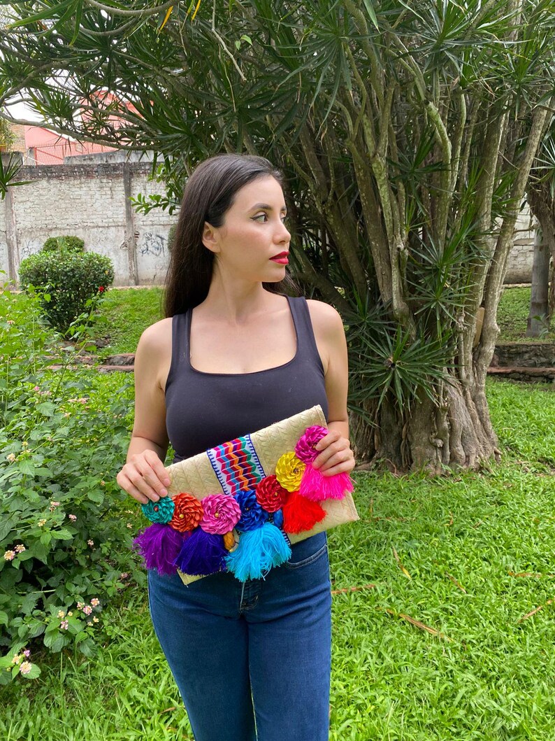 Handmade palm clutch featuring a rectangular woven palm leaf body, decorated with vibrant multicolor tassels and hand-painted pine cone flowers in pink, yellow, turquoise, purple, orange, and red. The center strip has a colorful embroidered textile accent, creating a unique artisan boho design. Perfect as a statement accessory for festivals, summer parties, vacations, or as a one-of-a-kind handmade gift for women.