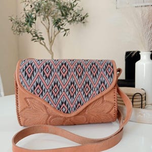 May include: A tan leather crossbody bag with a geometric patterned flap in pink, gray, and black. The bag features embossed floral detailing and a long, adjustable strap.