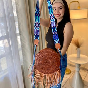 Handmade round leather bag with detailed embossed mandala design, long tan leather fringes, and vibrant handwoven multicolor strap in shades of blue and purple, showcased by a woman wearing jeans and a black top. The artisan Mexican purse rests elegantly in a modern indoor setting, ideal for boho chic fashion and handmade accessory lovers.