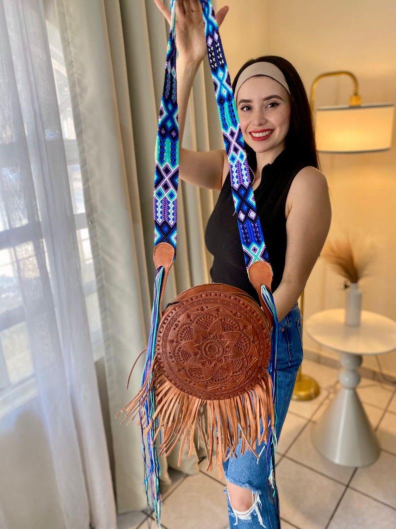 Handmade round leather bag with detailed embossed mandala design, long tan leather fringes, and vibrant handwoven multicolor strap in shades of blue and purple, showcased by a woman wearing jeans and a black top. The artisan Mexican purse rests elegantly in a modern indoor setting, ideal for boho chic fashion and handmade accessory lovers.