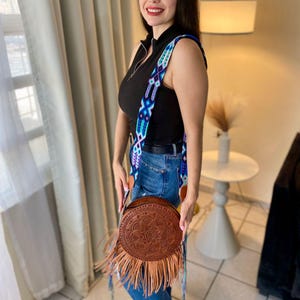 Handmade round leather bag with detailed embossed mandala design, long tan leather fringes, and vibrant handwoven multicolor strap in shades of blue and purple, showcased by a woman wearing jeans and a black top. The artisan Mexican purse rests elegantly in a modern indoor setting, ideal for boho chic fashion and handmade accessory lovers.