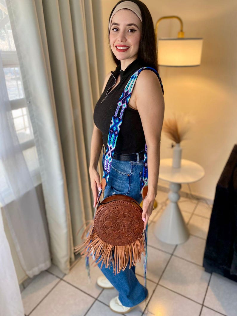 Handmade round leather bag with detailed embossed mandala design, long tan leather fringes, and vibrant handwoven multicolor strap in shades of blue and purple, showcased by a woman wearing jeans and a black top. The artisan Mexican purse rests elegantly in a modern indoor setting, ideal for boho chic fashion and handmade accessory lovers.