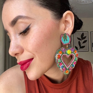 Pode incluir: Brincos em forma de coração com contas e um recorte em forma de coração. Os brincos apresentam uma mistura vibrante de cores, incluindo rosa, turquesa, amarelo e laranja, com um design floral. Eles são presos a um pequeno pino turquesa redondo.