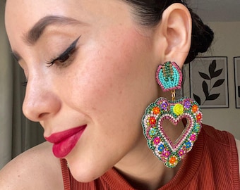 May include: Heart-shaped beaded earrings with a central heart cutout. The earrings feature a vibrant mix of colors, including pink, turquoise, yellow, and orange, with a floral design. The earrings are attached to a small, round turquoise stud.