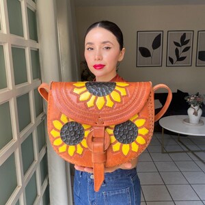 Handcrafted Mexican leather crossbody bag modeled by a woman in a rust sleeveless top and denim jeans. The bag is tan brown with hand-tooled sunflower details in bright yellow and black, featuring a curved flap, rustic stitching, and an adjustable leather strap. A unique bohemian accessory perfect for adding handmade floral charm and artisan craftsmanship to everyday outfits or travel looks.
