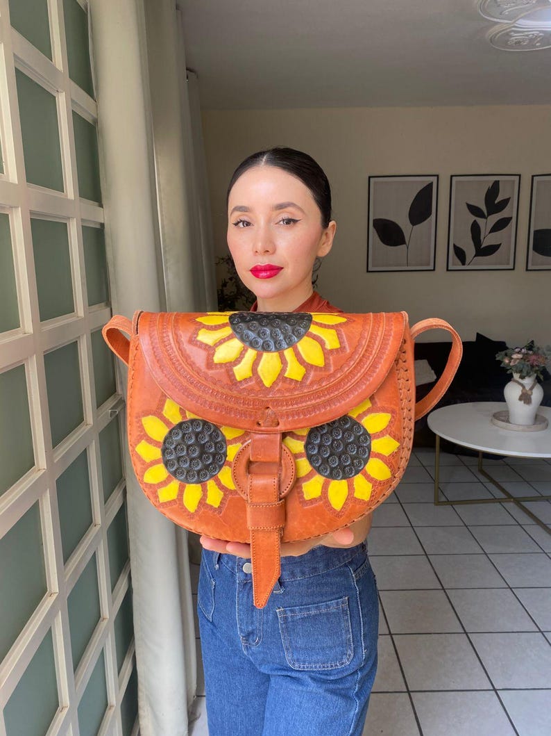 Handcrafted Mexican leather crossbody bag modeled by a woman in a rust sleeveless top and denim jeans. The bag is tan brown with hand-tooled sunflower details in bright yellow and black, featuring a curved flap, rustic stitching, and an adjustable leather strap. A unique bohemian accessory perfect for adding handmade floral charm and artisan craftsmanship to everyday outfits or travel looks.