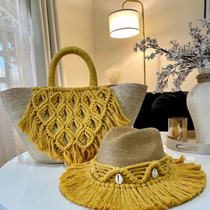 May include: A woven straw tote bag with a mustard yellow macrame design and fringe, paired with a matching fedora hat. The hat features a macrame band with seashells. The set is displayed on a white table, suggesting a beach or summer theme.