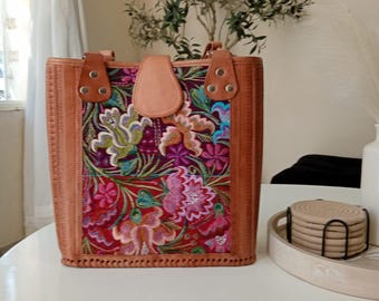 May include: A rectangular, tan leather tote bag with a colorful floral embroidered front panel. The bag has a top handle and a flap closure. The floral design includes pink, purple, blue, and green flowers on a red background.