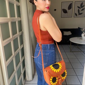 Handcrafted Mexican leather crossbody bag modeled by a woman in a rust sleeveless top and denim jeans. The bag is tan brown with hand-tooled sunflower details in bright yellow and black, featuring a curved flap, rustic stitching, and an adjustable leather strap. A unique bohemian accessory perfect for adding handmade floral charm and artisan craftsmanship to everyday outfits or travel looks.