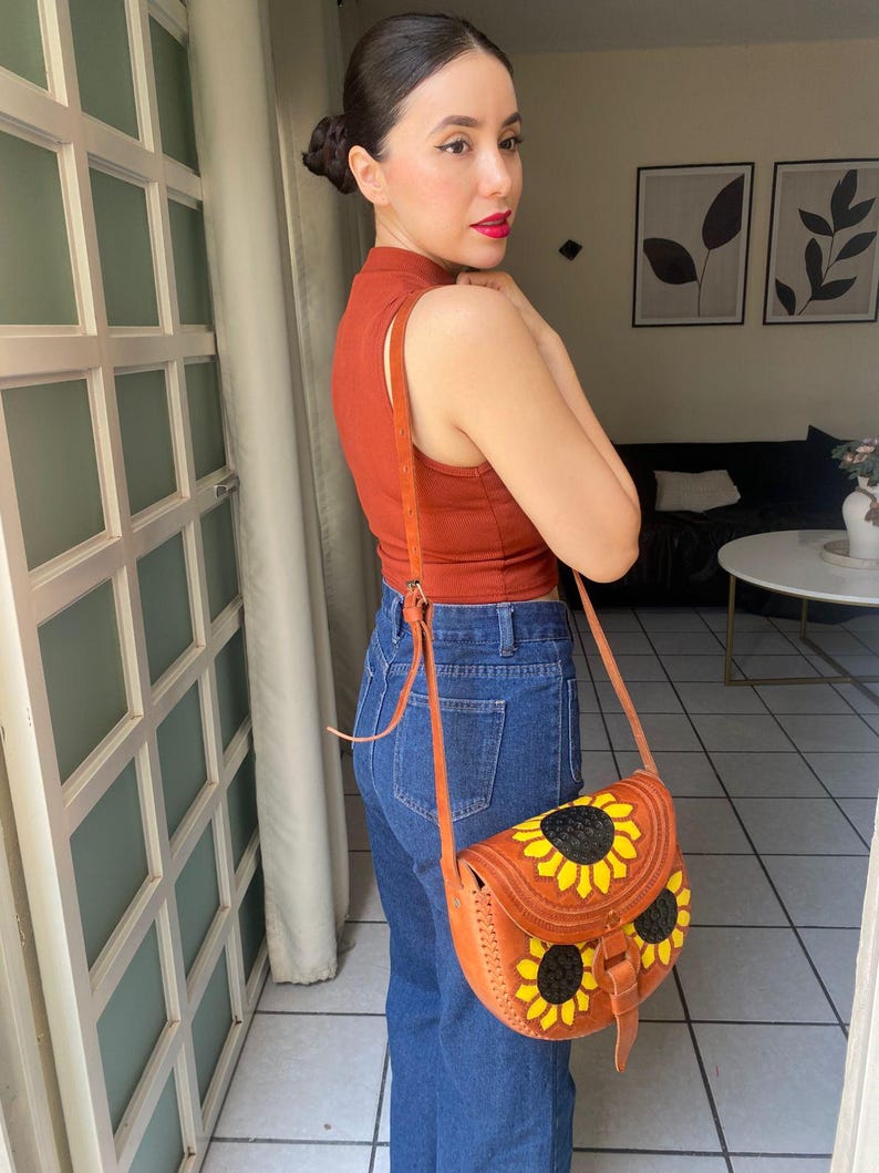 Handcrafted Mexican leather crossbody bag modeled by a woman in a rust sleeveless top and denim jeans. The bag is tan brown with hand-tooled sunflower details in bright yellow and black, featuring a curved flap, rustic stitching, and an adjustable leather strap. A unique bohemian accessory perfect for adding handmade floral charm and artisan craftsmanship to everyday outfits or travel looks.