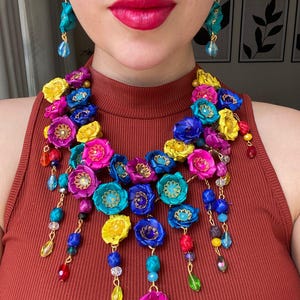 Pode incluir: Um vibrante colar floral e brincos a condizer. O colar apresenta flores de tecido coloridas em tons de rosa, azul e amarelo, acentuadas com contas penduradas. Os brincos espelham o design floral e de contas.