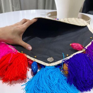 Handmade palm clutch featuring a rectangular woven palm leaf body, decorated with vibrant multicolor tassels and hand-painted pine cone flowers in pink, yellow, turquoise, purple, orange, and red. The center strip has a colorful embroidered textile accent, creating a unique artisan boho design. Perfect as a statement accessory for festivals, summer parties, vacations, or as a one-of-a-kind handmade gift for women.