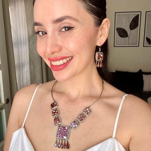 Op de afbeelding: Een zilveren ketting met een rode en witte gepatenteerde hanger en bijpassende oorbellen. De hanger is vierkant met een kleiner vierkant in het midden. De oorbellen zijn ook vierkant met een kleiner vierkant in het midden. De ketting en oorbellen zijn gemaakt van zilver en hebben een rood en wit gepatenteerd ontwerp.