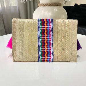Handmade palm clutch featuring a rectangular woven palm leaf body, decorated with vibrant multicolor tassels and hand-painted pine cone flowers in pink, yellow, turquoise, purple, orange, and red. The center strip has a colorful embroidered textile accent, creating a unique artisan boho design. Perfect as a statement accessory for festivals, summer parties, vacations, or as a one-of-a-kind handmade gift for women.