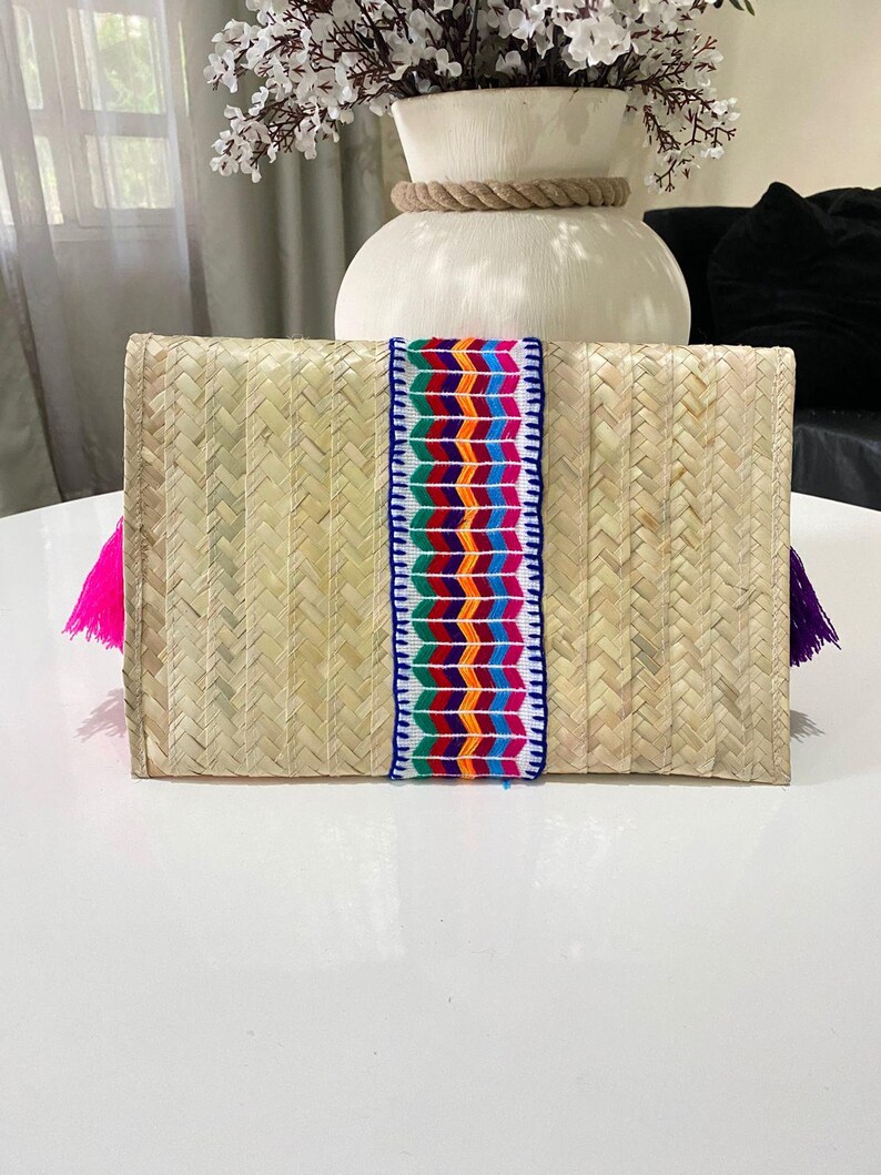 Handmade palm clutch featuring a rectangular woven palm leaf body, decorated with vibrant multicolor tassels and hand-painted pine cone flowers in pink, yellow, turquoise, purple, orange, and red. The center strip has a colorful embroidered textile accent, creating a unique artisan boho design. Perfect as a statement accessory for festivals, summer parties, vacations, or as a one-of-a-kind handmade gift for women.