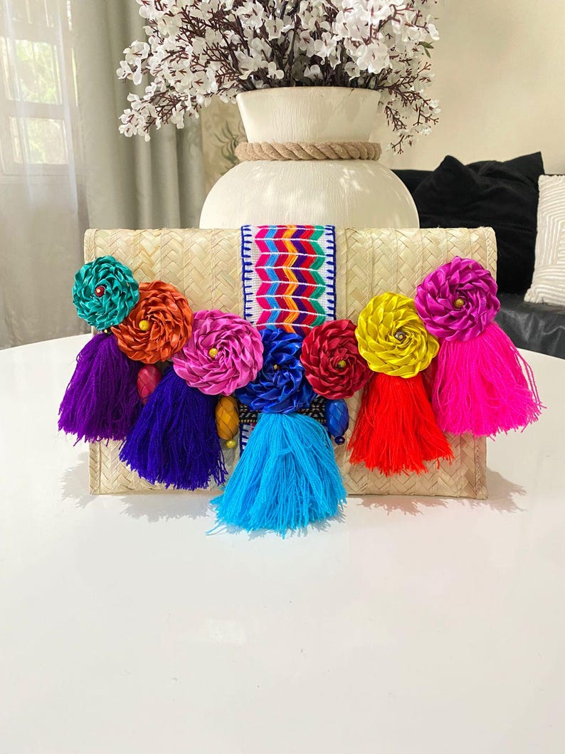 Handmade palm clutch featuring a rectangular woven palm leaf body, decorated with vibrant multicolor tassels and hand-painted pine cone flowers in pink, yellow, turquoise, purple, orange, and red. The center strip has a colorful embroidered textile accent, creating a unique artisan boho design. Perfect as a statement accessory for festivals, summer parties, vacations, or as a one-of-a-kind handmade gift for women.