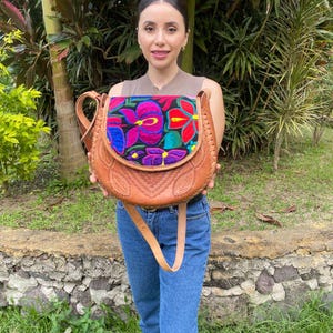Könnte beinhalten: Braune Leder-Umhängetasche mit Klappenverschluss. Die Klappe hat ein lebendiges, gesticktes Blumendesign in Pink-, Blau- und Grüntönen. Die Tasche hat einen langen, verstellbaren Riemen und wird von einer Person gehalten.
