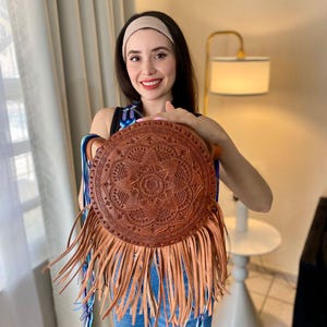 Handmade round leather bag with detailed embossed mandala design, long tan leather fringes, and vibrant handwoven multicolor strap in shades of blue and purple, showcased by a woman wearing jeans and a black top. The artisan Mexican purse rests elegantly in a modern indoor setting, ideal for boho chic fashion and handmade accessory lovers.