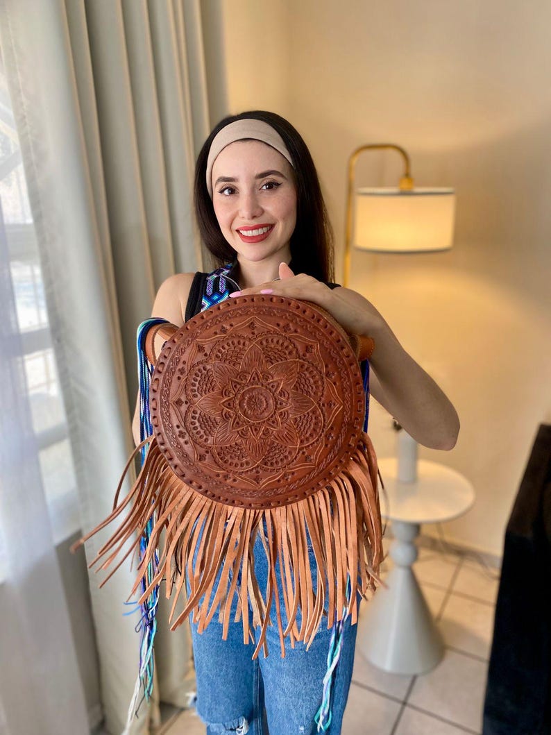Handmade round leather bag with detailed embossed mandala design, long tan leather fringes, and vibrant handwoven multicolor strap in shades of blue and purple, showcased by a woman wearing jeans and a black top. The artisan Mexican purse rests elegantly in a modern indoor setting, ideal for boho chic fashion and handmade accessory lovers.