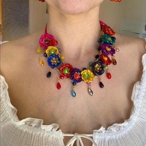 Könnte beinhalten: Eine bunte Blumenkette mit roten, gelben, blauen, grünen und violetten Blüten. Die Kette besteht aus Stoffblumen und goldenen Perlen. Die Kette hängt an einem Hals.