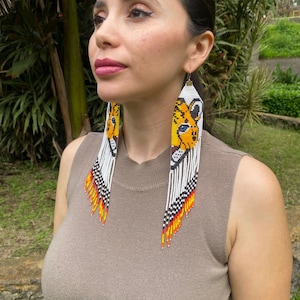 May include: Long, beaded earrings featuring a yellow and black animal face design. The earrings have a white background with black, red, and yellow fringe. The earrings are worn by a person wearing a brown sleeveless top.
