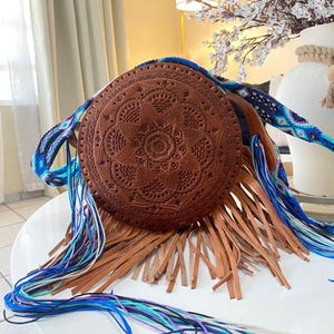 Handmade round leather bag with detailed embossed mandala design, long tan leather fringes, and vibrant handwoven multicolor strap in shades of blue and purple, showcased by a woman wearing jeans and a black top. The artisan Mexican purse rests elegantly in a modern indoor setting, ideal for boho chic fashion and handmade accessory lovers.