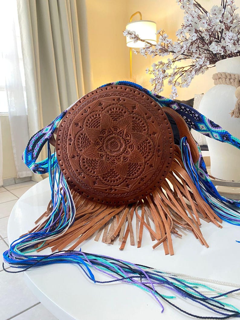 Handmade round leather bag with detailed embossed mandala design, long tan leather fringes, and vibrant handwoven multicolor strap in shades of blue and purple, showcased by a woman wearing jeans and a black top. The artisan Mexican purse rests elegantly in a modern indoor setting, ideal for boho chic fashion and handmade accessory lovers.