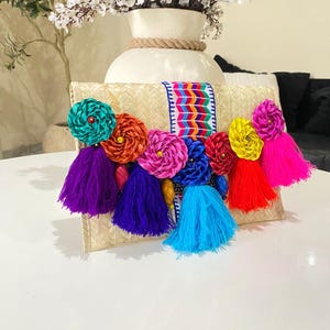 Handmade palm clutch featuring a rectangular woven palm leaf body, decorated with vibrant multicolor tassels and hand-painted pine cone flowers in pink, yellow, turquoise, purple, orange, and red. The center strip has a colorful embroidered textile accent, creating a unique artisan boho design. Perfect as a statement accessory for festivals, summer parties, vacations, or as a one-of-a-kind handmade gift for women.