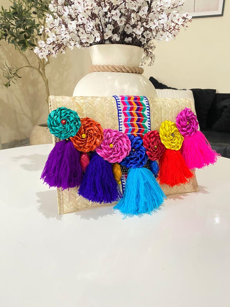 Handmade palm clutch featuring a rectangular woven palm leaf body, decorated with vibrant multicolor tassels and hand-painted pine cone flowers in pink, yellow, turquoise, purple, orange, and red. The center strip has a colorful embroidered textile accent, creating a unique artisan boho design. Perfect as a statement accessory for festivals, summer parties, vacations, or as a one-of-a-kind handmade gift for women.