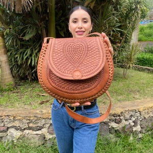 May include: A brown leather crossbody bag with a woven design on the flap and body. The bag has a long strap and a circular clasp. The bag is held by a person wearing blue jeans.