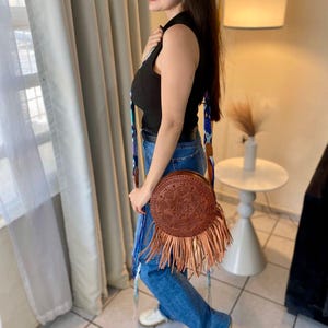 Handmade round leather bag with detailed embossed mandala design, long tan leather fringes, and vibrant handwoven multicolor strap in shades of blue and purple, showcased by a woman wearing jeans and a black top. The artisan Mexican purse rests elegantly in a modern indoor setting, ideal for boho chic fashion and handmade accessory lovers.