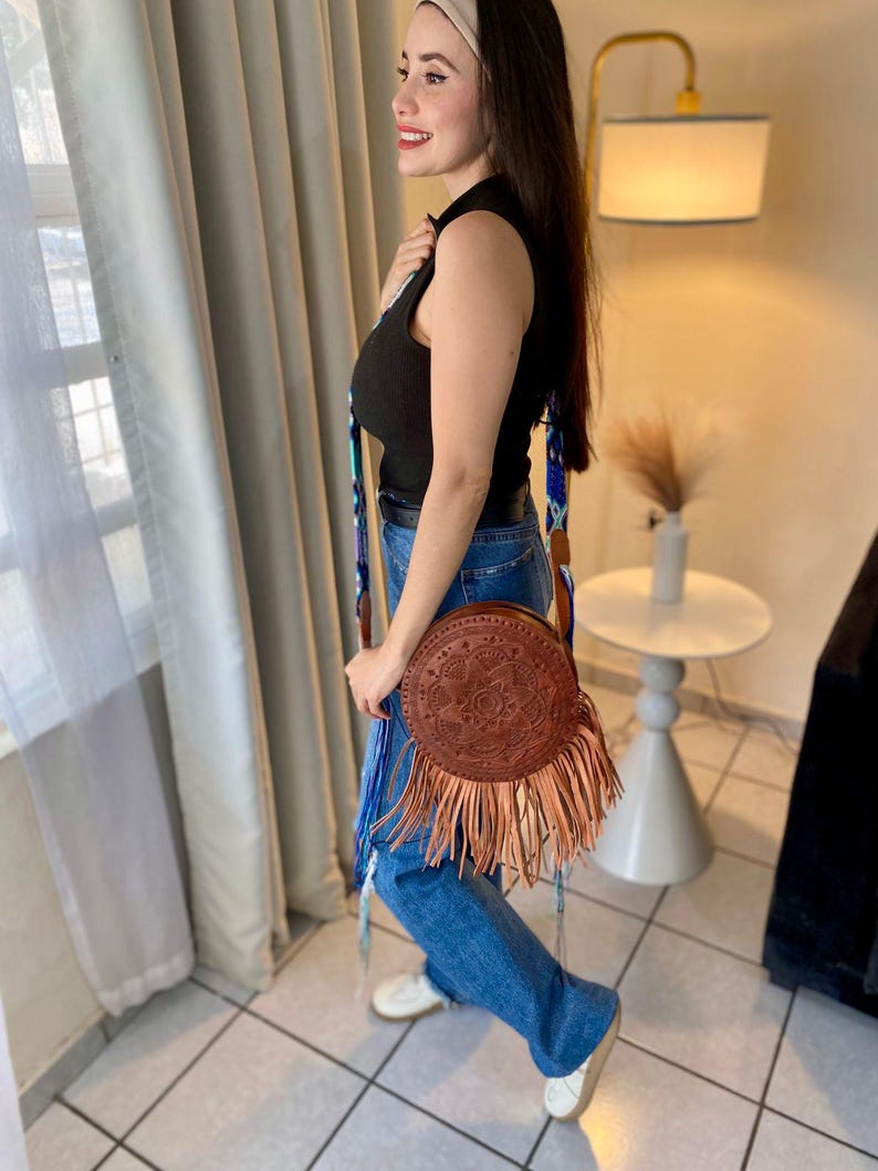 Handmade round leather bag with detailed embossed mandala design, long tan leather fringes, and vibrant handwoven multicolor strap in shades of blue and purple, showcased by a woman wearing jeans and a black top. The artisan Mexican purse rests elegantly in a modern indoor setting, ideal for boho chic fashion and handmade accessory lovers.