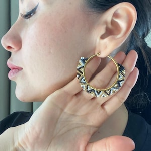 May include: Gold hoop earrings with a beaded design. The earrings have a gold hoop with a section covered in black, white, and gold beads. The earrings are held up to the ear.