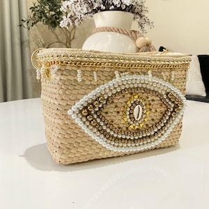 May include: Woven straw handbag with a decorative eye design. The bag features a rectangular shape, adorned with pearls and gold beads. A small seashell is at the center of the eye. The top edge is trimmed with gold chain and pearls.
