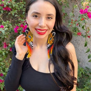 Può includere: Una donna indossa una camicia nera a maniche lunghe e jeans blu con capelli lunghi e neri. Indossa lunghi orecchini a perline con un design di giaguaro in nero, giallo e arancione.