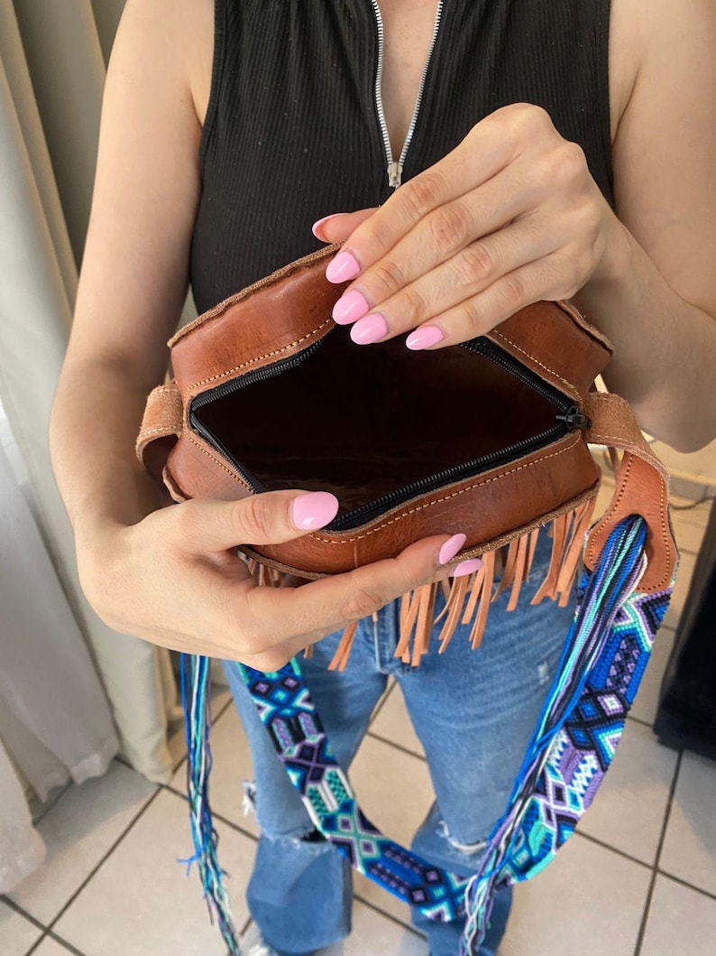 Handmade round leather bag with detailed embossed mandala design, long tan leather fringes, and vibrant handwoven multicolor strap in shades of blue and purple, showcased by a woman wearing jeans and a black top. The artisan Mexican purse rests elegantly in a modern indoor setting, ideal for boho chic fashion and handmade accessory lovers.