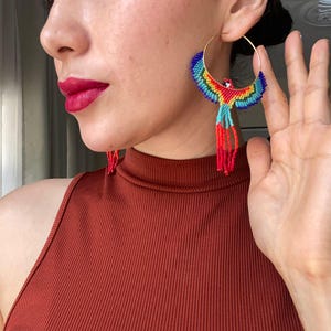 Puede incluir: Pendientes de aro dorados con un diseño de loro de cuentas. El loro presenta colores vibrantes que incluyen azul, rojo, amarillo y verde. Los pendientes tienen largas borlas de cuentas rojas.