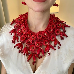 Peut inclure: Un collier floral rouge vif et des boucles d'oreilles assorties. Le collier présente de nombreuses petites fleurs rouges détaillées avec des accents dorés et des breloques rouges en forme de pétales. Les boucles d'oreilles reprennent le motif floral. Le bijou est porté sur un haut blanc.