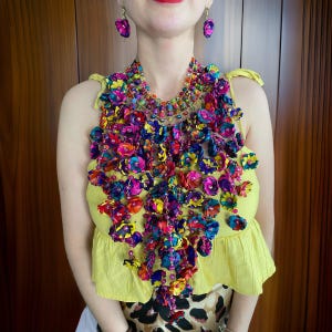 Könnte beinhalten: Eine farbenfrohe Statement-Halskette aus Stoffblumen in den Farben Pink, Lila, Gelb und Grün. Die Kette ist mit Perlen besetzt und hängt bis zur Brust.