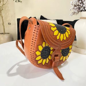 Handcrafted Mexican leather crossbody bag modeled by a woman in a rust sleeveless top and denim jeans. The bag is tan brown with hand-tooled sunflower details in bright yellow and black, featuring a curved flap, rustic stitching, and an adjustable leather strap. A unique bohemian accessory perfect for adding handmade floral charm and artisan craftsmanship to everyday outfits or travel looks.